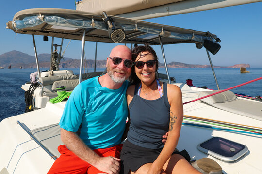 A smiling couple relaxes on a sailboat deck surrounded by calm blue water and volcanic islands near Milos, enjoying Mediterranean sunshine and coastal adventure.