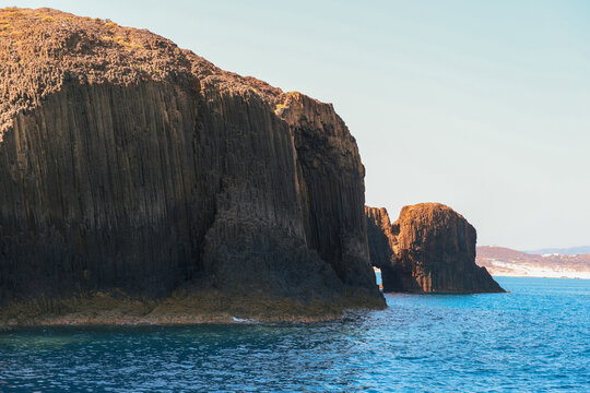 Massive columnar basalt cliffs rise from turquoise water along the coast of Milos, showcasing dramatic volcanic geology and the island&rsquo;s rugged Mediterranean beauty.