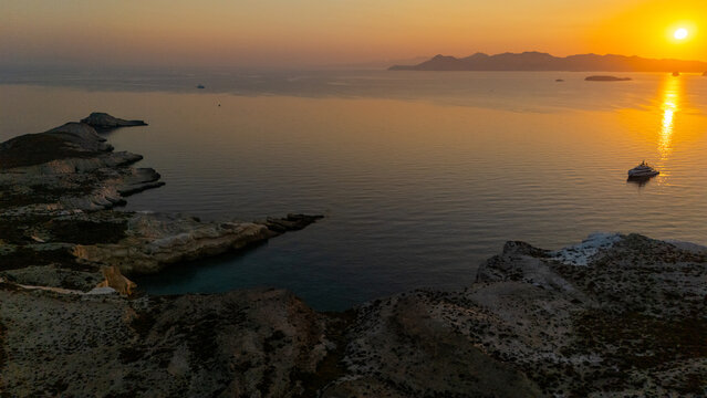 Golden sunlight rises over the rocky coastline of Mandrakia on Milos, reflecting across calm water and illuminating the rugged cliffs of the Aegean in early morning light.