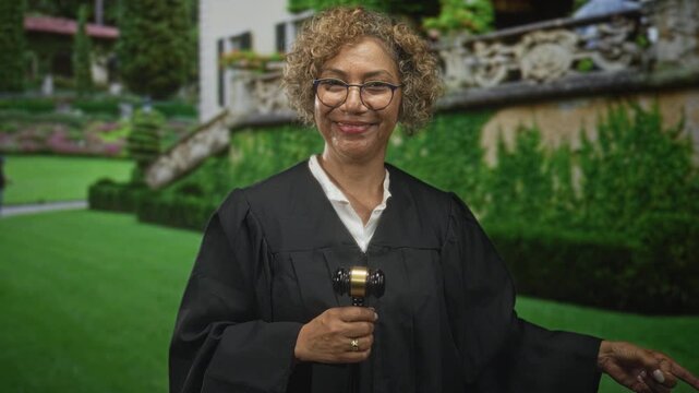 Middleaged woman judge in robe holds gavel and points finger by building balustrade and manicured lawn, smiling with assertive posture; confidence justice dignity.