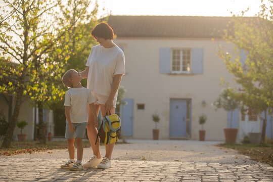 mother and child standing together on a cobblestone path in a warm and familial village setting with white building, blue doors, and potted plants, eye-level perspective.