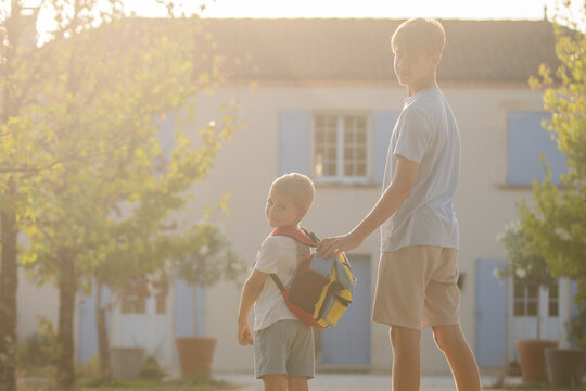 Two boys walking together, in outdoor residential area with trees and buildings in, with school backpack, trees, buildings, from a side view of the boys perspective.