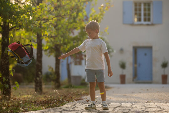 child standing on cobblestone path in residential area with calm mood and red backpack, side view, white shirt, gray shorts, family portraits, childhood memories.