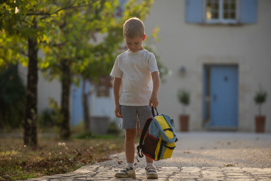 young boy standing with backpack on outdoor pathway near building, calm side view, cotton and plastic materials, white and blue colors, school setting, childhood education.