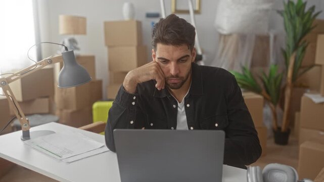 Man at laptop with hand on chin amid stacked cardboard moving boxes and desk lamp in a residential building; pensive transition.