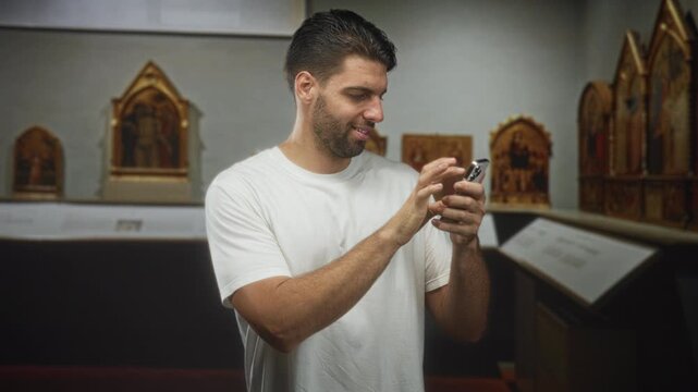 Hispanic man with beard holding smartphone, tapping screen and pumping fist in building museum gallery with framed altarpieces visible; casual celebration joy.