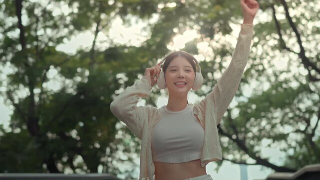 Happy young Asian woman dancing to music with wireless headphones outdoors, joyful active lifestyle and entertainment concept