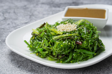 Fresh hiyashi wakame salad with sesame seeds and peanut dressing against grey concrete background. Green seaweed chuka salad on a white plate with sauce bowl, close up view with copy space. © 0990930308
