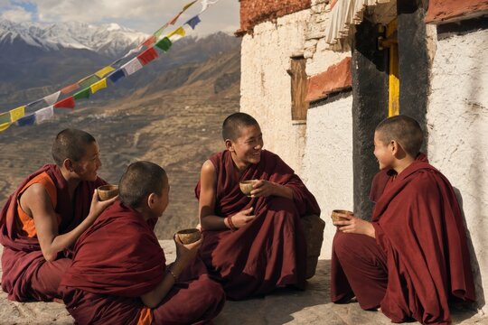 Buddhist monks sharing tea outside a mountain monastery. Spiritual community and traditional Himalayan culture with prayer flags, ancient architecture, and snowy mountain landscape.