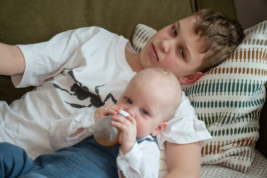 Older brother cuddles baby sibling while younger child drinks from bottle resting safely in his arms on sofa at home. Sibling care, brotherhood, caregiving, responsibility, family bond, support, child