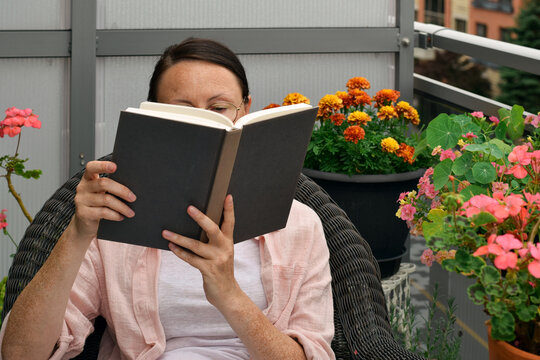 Middle aged woman sitting in wicker chair reading hardcover book with potted flowers in background on balcony.