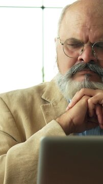 Indian Asian elderly businessman with a beard, dressed in a coat, seated in a modern office, thoughtfully using a laptop, focusing on business work