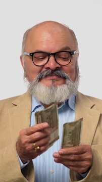 Indian Asian elderly businessman with a beard and suit, standing against a white background counting Indian currency notes ,enjoying shower of  money and  celebrating success by gesture of fist