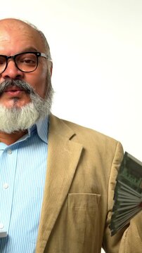 Indian Asian senior businessman with a beard, wearing a suit, stands against a white background, holding a fan of money in one hand and a laptop in the other, smiling happily 