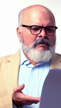 An Indian Asian elderly businessman with a beard is standing against a white background,wearing a suit  looking through important business documents,reviewing with  disappointed expression
