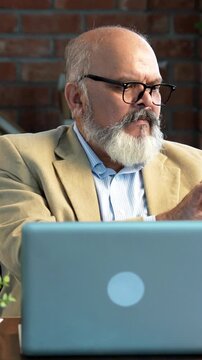 ndian Asian elderly businessman with a beard, wearing a suit, sitting in a modern office setup, working on a laptop and using a calculator to check or review financial transactions