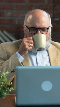 Indian Asian elderly businessman with a beard and suit sitting in a modern office setup, attending a video call on a laptop, holding documents in hand, and sipping coffee, showing multitasking 