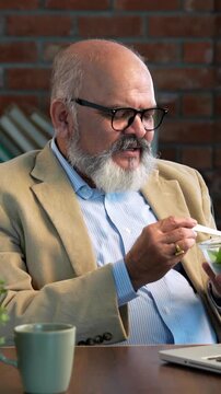 Indian Asian elderly businessman sporting a beard in  a suit, attending a video call on his laptop in modern set up while enjoying a midday snack of salad and coffee, balancing work and health 