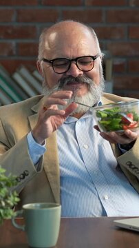 Indian Asian elderly businessman with a beard, wearing a suit, sitting in a beautiful modern office, working on a laptop while enjoying a midday snack of salad and  coffee