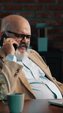 Indian Asian elderly businessman with a beard, wearing a suit, sitting in a beautiful modern office, attending a phone call while working on his laptop
