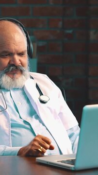 Indian Asian elderly doctor with a beard, wearing an apron and stethoscope, is conducting an online consultation on his laptop, in modern healthcare setup