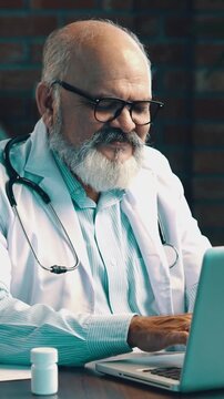 Indian Asian elderly doctor with beard, wearing white coat and stethoscope, giving thumbs up while looking happy during online consultation on laptop, reading test reports at modern clinic