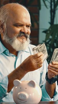 Indian Asian elderly businessman with beard counting Indian money with a smile, coins and piggy bank on table, happily showing his satisfaction in saving money