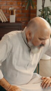 Indian Asian senior businessman with beard, sitting on sofa at night, using cellphone, showing pain from backache, in a modern home setting