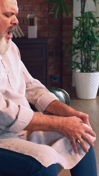 Indian Asian elderly businessman with a beard, sitting on a sofa in a modern home, tries to exercise his knee by folding his leg  ,shows severe pain on his face 