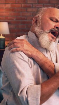 Indian Asian elderly businessman with a beard, sitting on a sofa in a modern home, experiencing severe shoulder pain showing a painful expression on his face