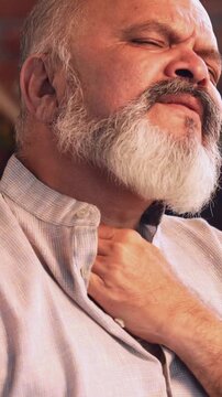 Indian Asian elderly businessman with a beard sitting on a sofa in a modern home, suffering from severe neck pain, trying to massage and move his neck while showing a painful expression on his face