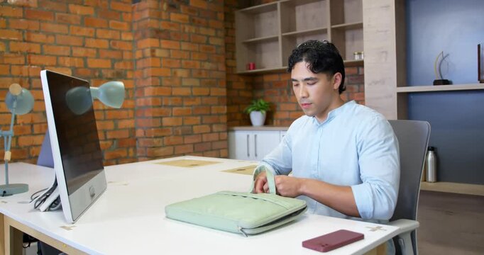 Adult Asian male in blue shirt at desk, seeing green laptop sleeve, unzipping, preparing laptop