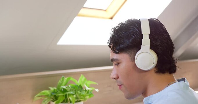 Asian man sitting under skylight, watching laptop, listening to laptop on headphones, copy space