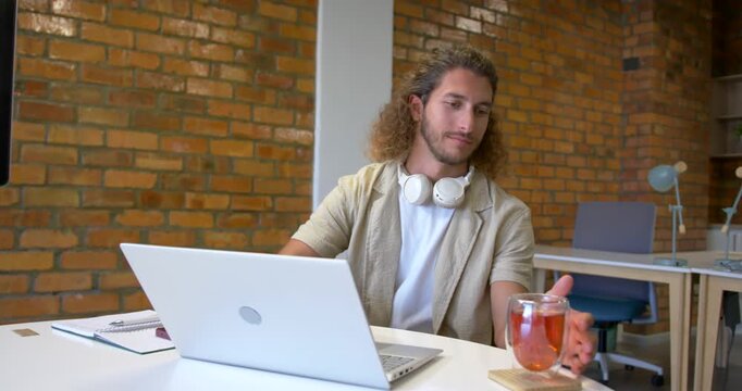 Typing on laptop, man glancing at mug, adjusting coaster, sipping tea for refreshment at white desk