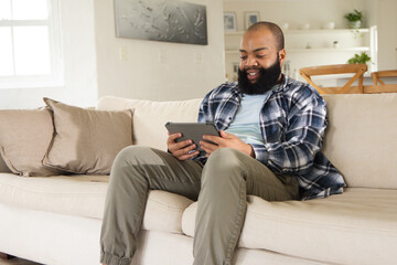 Fototapeta premium Adult African American man sitting on beige sofa in living room holding tablet, wearing plaid shirt