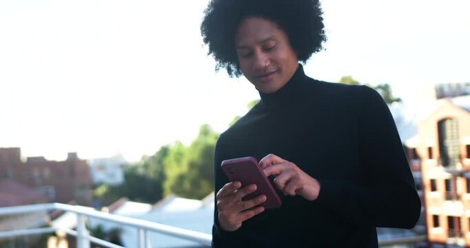 African American man standing on balcony in black turtleneck holding pink phone tapping, scrolling