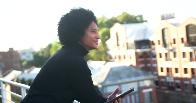 African American man leaning on roof railing with tablet, glancing at tablet, lifting chin, pausing