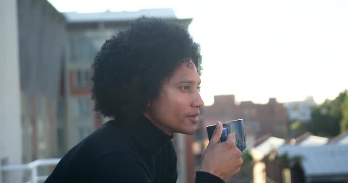 African American man sitting on balcony turtleneck gazing city raising blue ceramic mug sipping