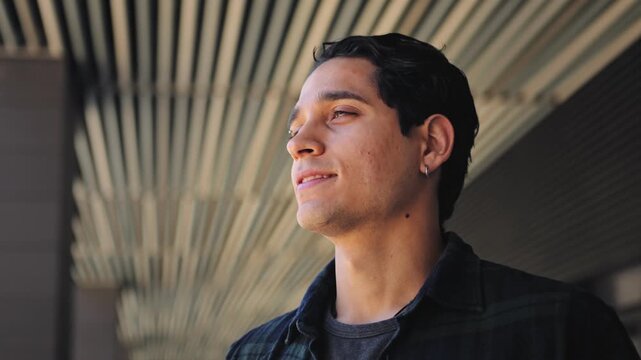 Relaxed young hispanic man looking away outdoors near modern office building in urban city