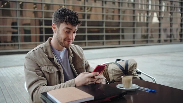 Young man using mobile phone and drinking coffee at a table with laptop and notebook after work break