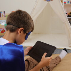 Back view of a child wearing a blue superhero cape sitting on the floor holding a screen tablet near a teepee tent © Imageologist