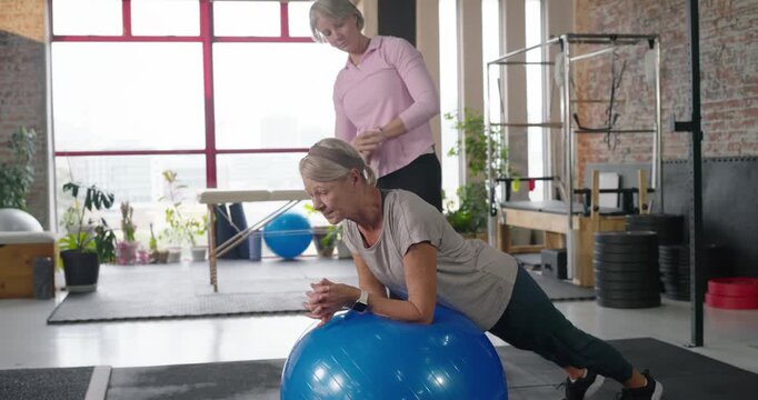 Female trainer approaching, adjusting client holding plank on blue Swiss ball for form check at gym