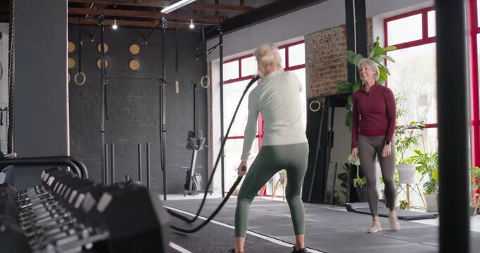 Female partners training in gym by dumbbells, rope user grasping battle ropes and making waves