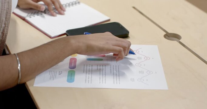 Indian woman opening notebook and tracing infographic with blue pen at table, reviewing for meeting