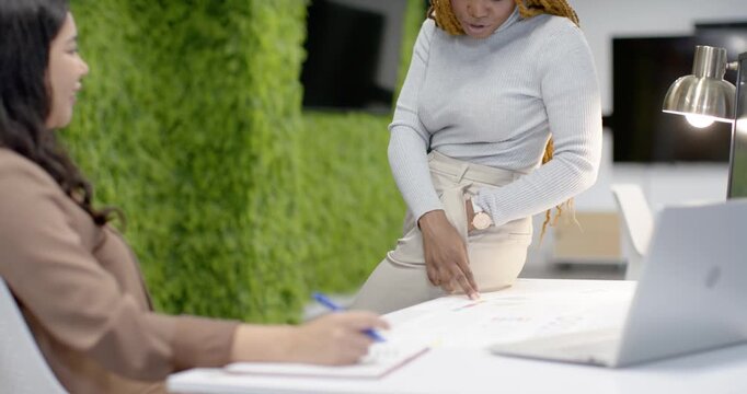 Diverse female team leaning and pointing at charts in office taking notes using PC reviewing data