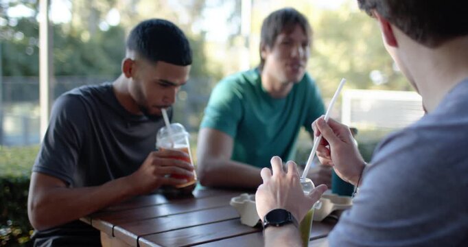 Diverse male friends noticing drink cups, sitting at picnic table handling straws and chatting