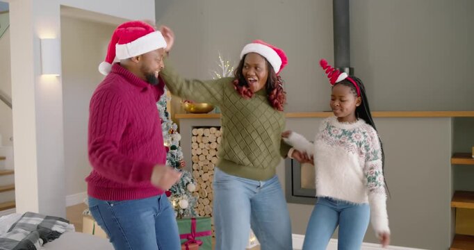 Family wearing Santa hats moving to music, solo steps joining arms, dancing and high-fiving by tree