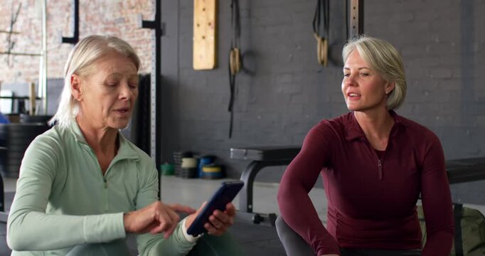 Senior and adult women in zip tops tapping smartphone, rotating screen, causing smiles on gym mats