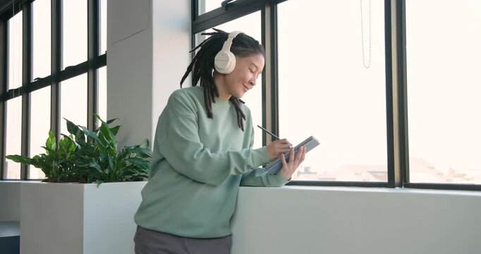 African American nonbinary in mint top entering left, tapping stylus on tablet by window for ideas