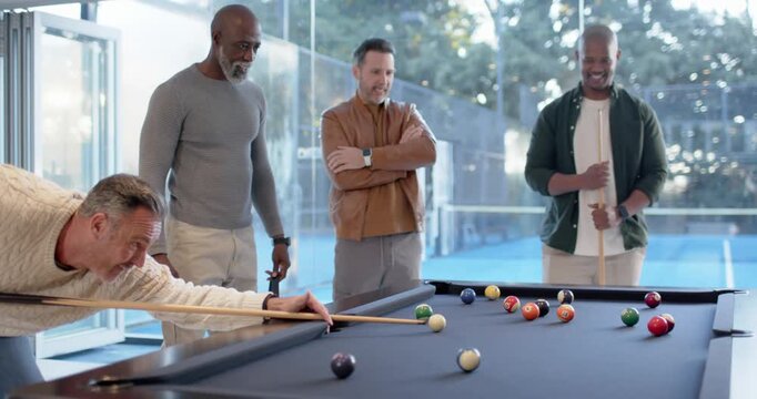 Diverse male friends playing pool at slate-blue table, shooter striking cue ball, balls scattering